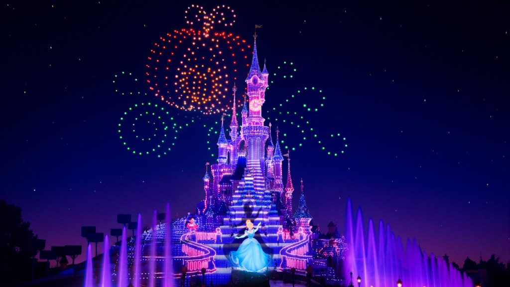 Drone lights in shape of pumpkin in sky behind castle of Sleeping Beauty in Disneyland Paris, with a projection of Cinderella.